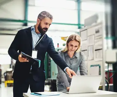 Interim Management Operations: Das Bild zeigt einen Operations-Manager, der seiner Kollegin etwas auf einem Laptop zeigt.